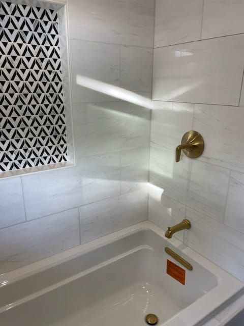 White tiled bathtub and shower area with gold fixtures and patterned wall insert.