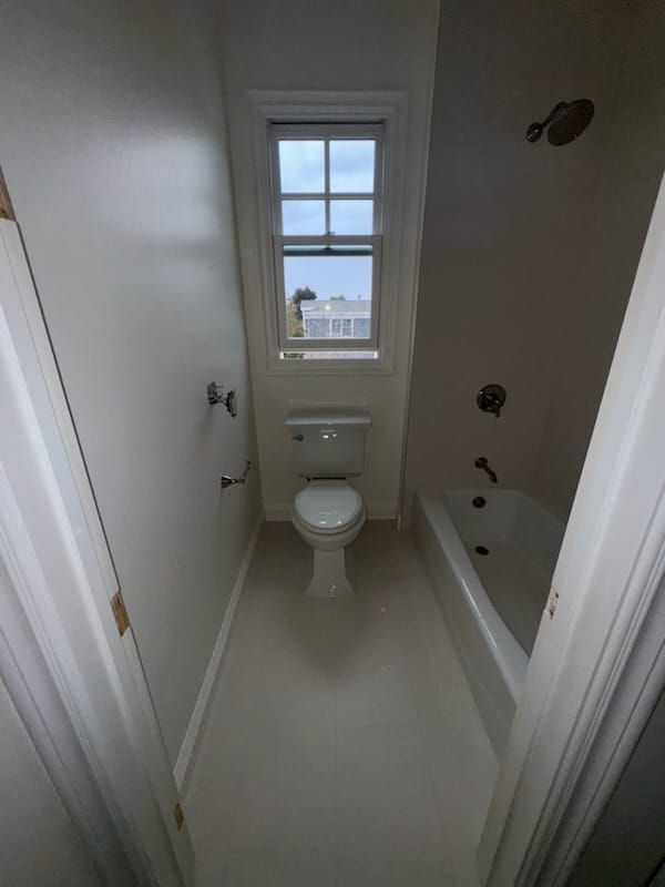 Narrow bathroom with a toilet, bathtub, and small window. Off-white walls and floor.