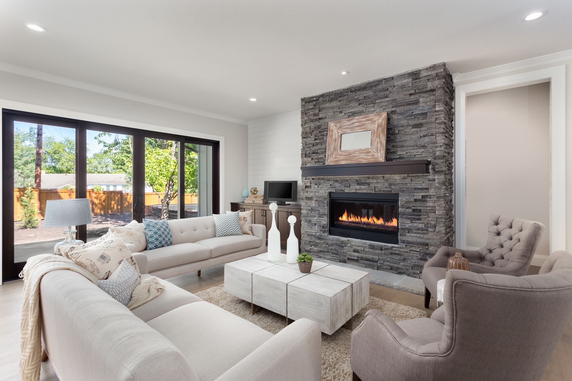 Bright modern living room with white sofas, stone fireplace, and large sliding glass doors to a patio.