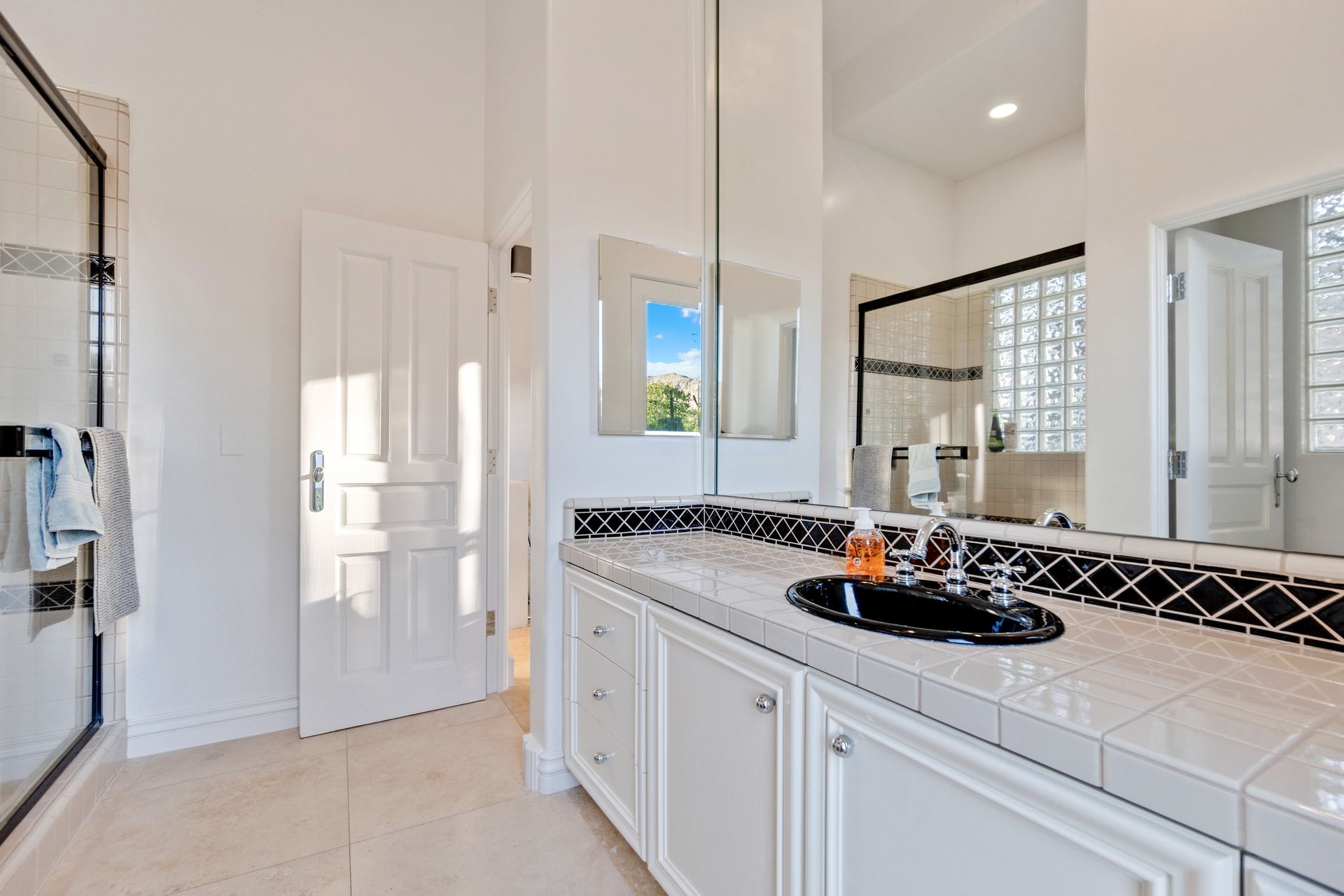Bright bathroom with white cabinets, tiled vanity, glass shower, and a black countertop with toiletries.