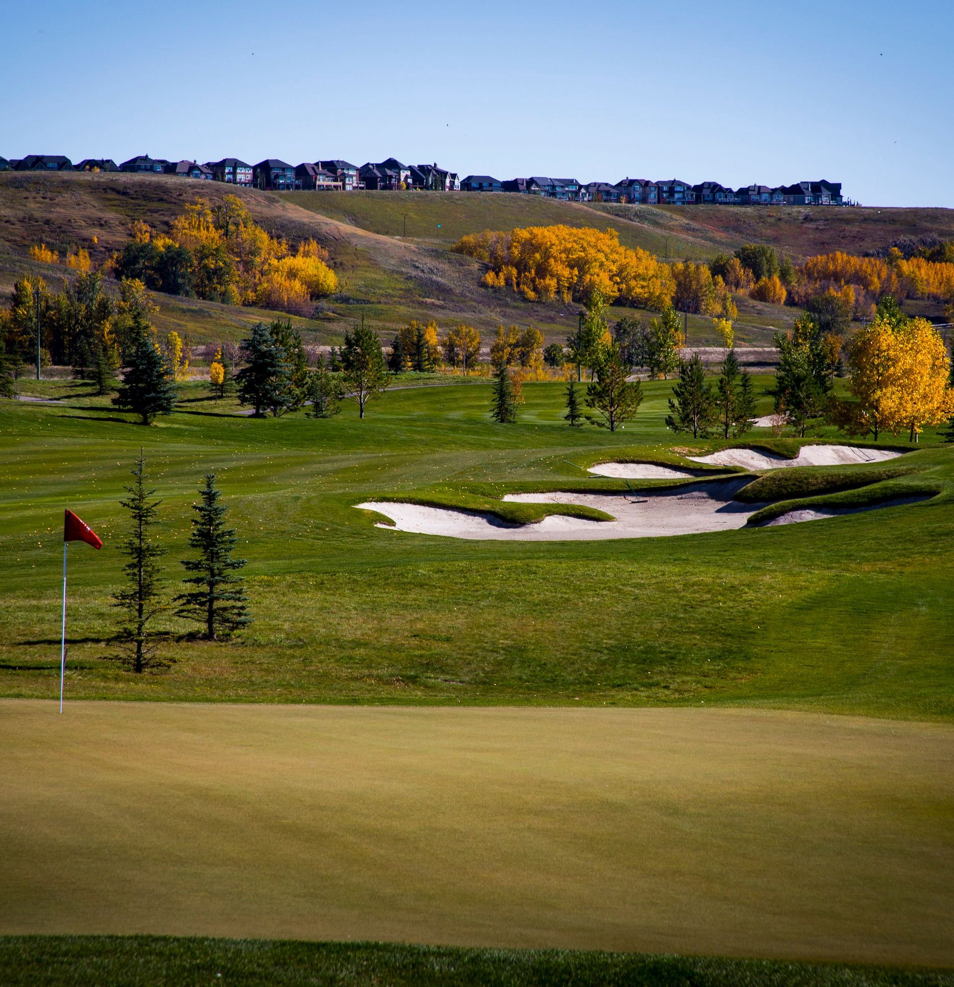 Golf course green with red flag. Sand traps and trees line the fairway. Houses on a hill in the background.