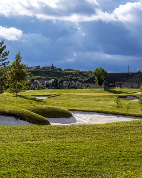 Golf course with green grass, sand traps, and a cloudy sky. Houses and a building are in the distance.