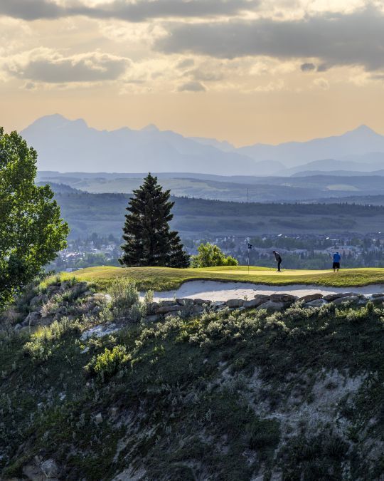 Two golfers on a scenic clifftop putting green with a sand bunker, pine tree, and distant mountains under a cloudy sky.