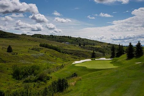 Green golf course with trees, bunkers, and houses on a hill against a blue sky with clouds.