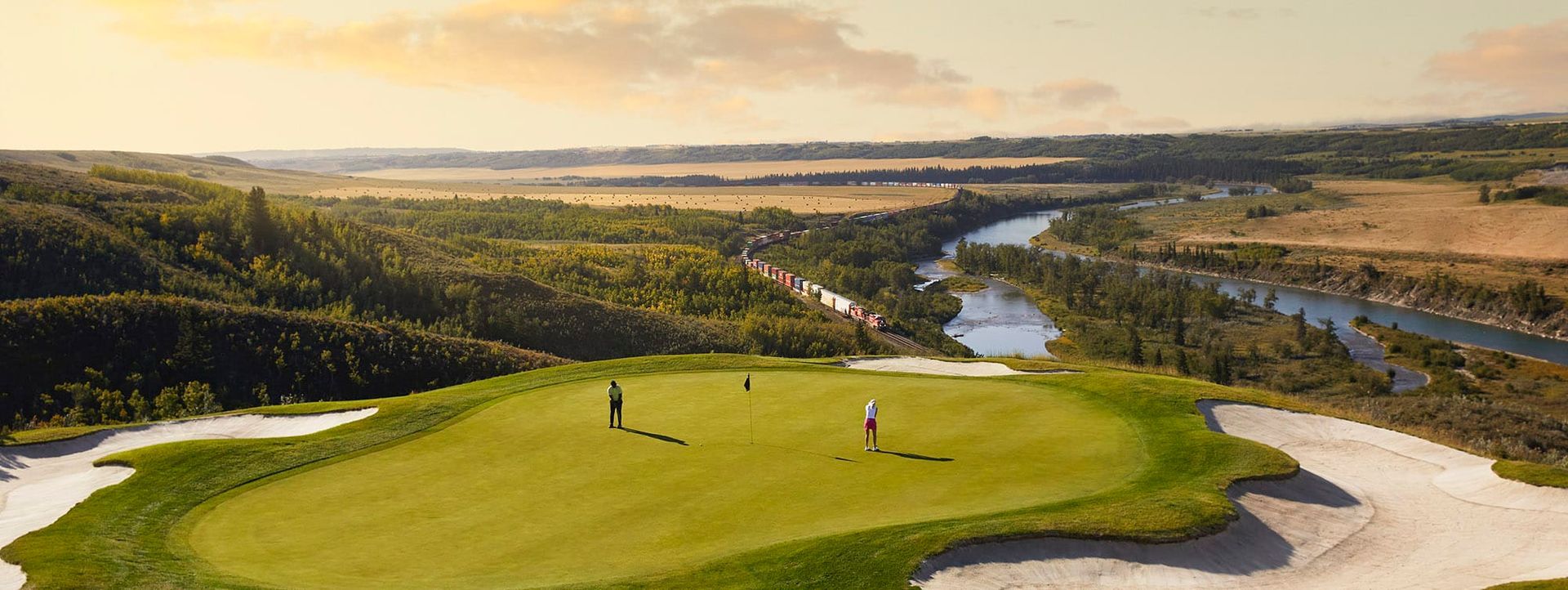 Golfers on a green course overlooking a river and valley in warm sunlight.
