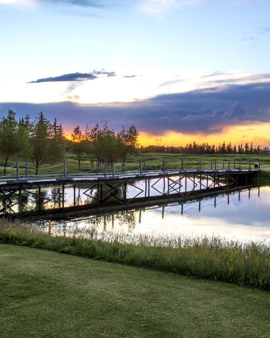Wooden bridge over a calm pond, reflecting the sunset sky; green grass in foreground.