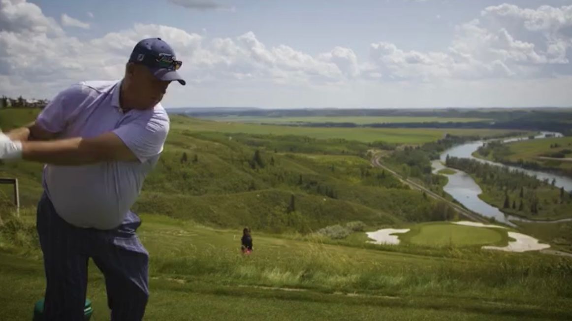 Golfer in blue shirt swings club on golf course overlooking a river and green landscape.