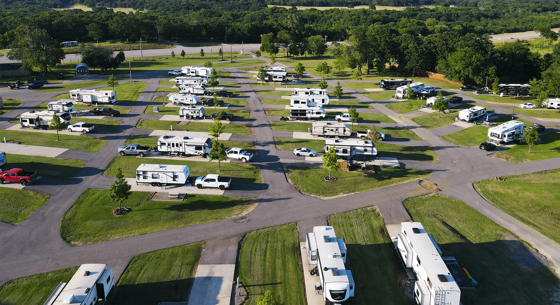 Aerial view of a campground with rows of white RVs and trailers on paved lots surrounded by green trees.
