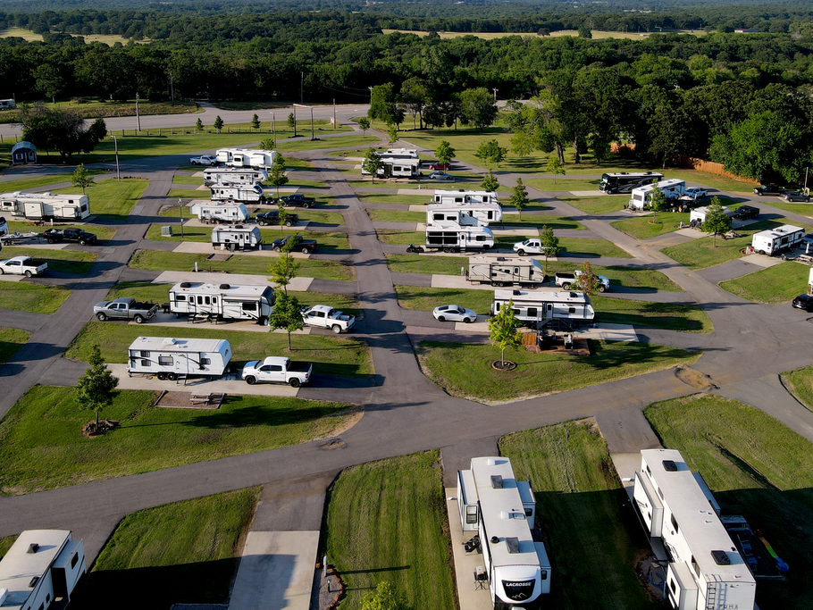 Aerial view of a grassy RV park with parked campers, trailers, and cars along paved lanes.