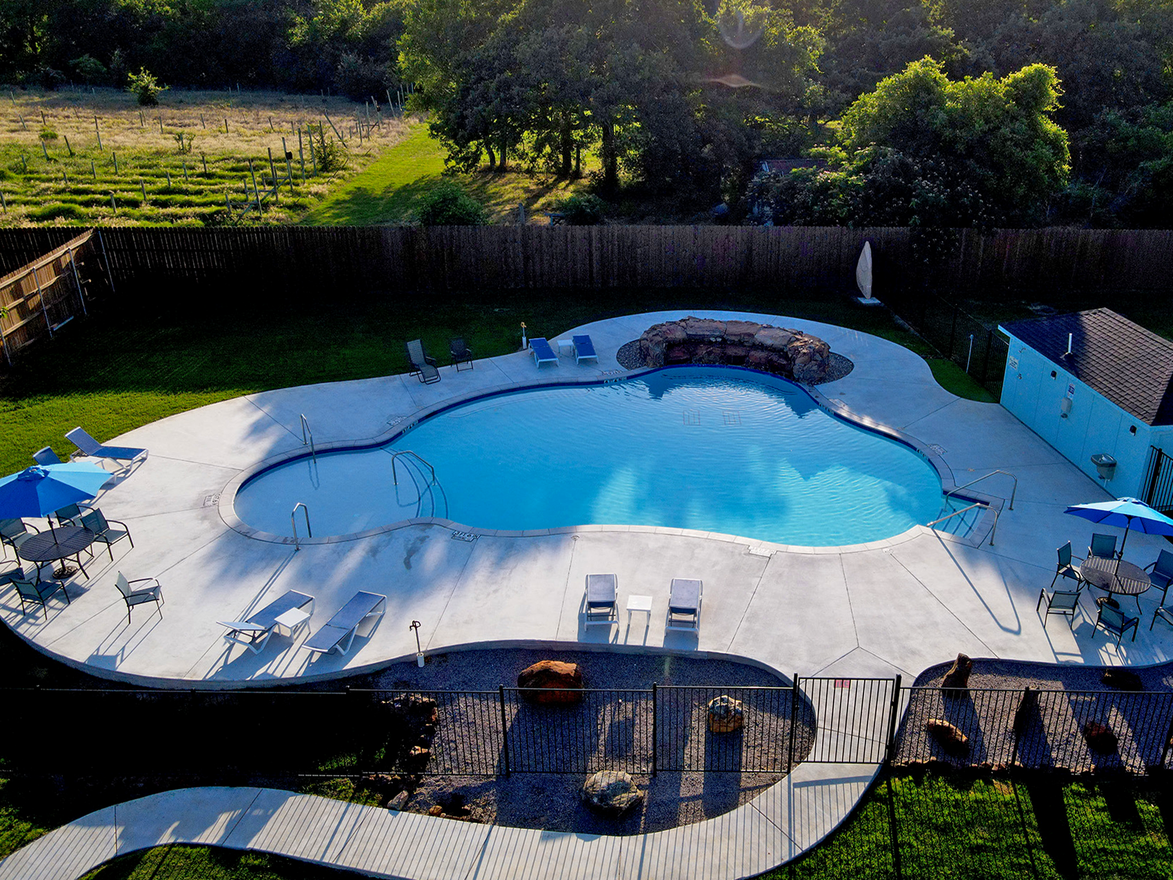 RV park swimming pool with a white deck, blue water, and stone waterfall, bordered by trees and lawn