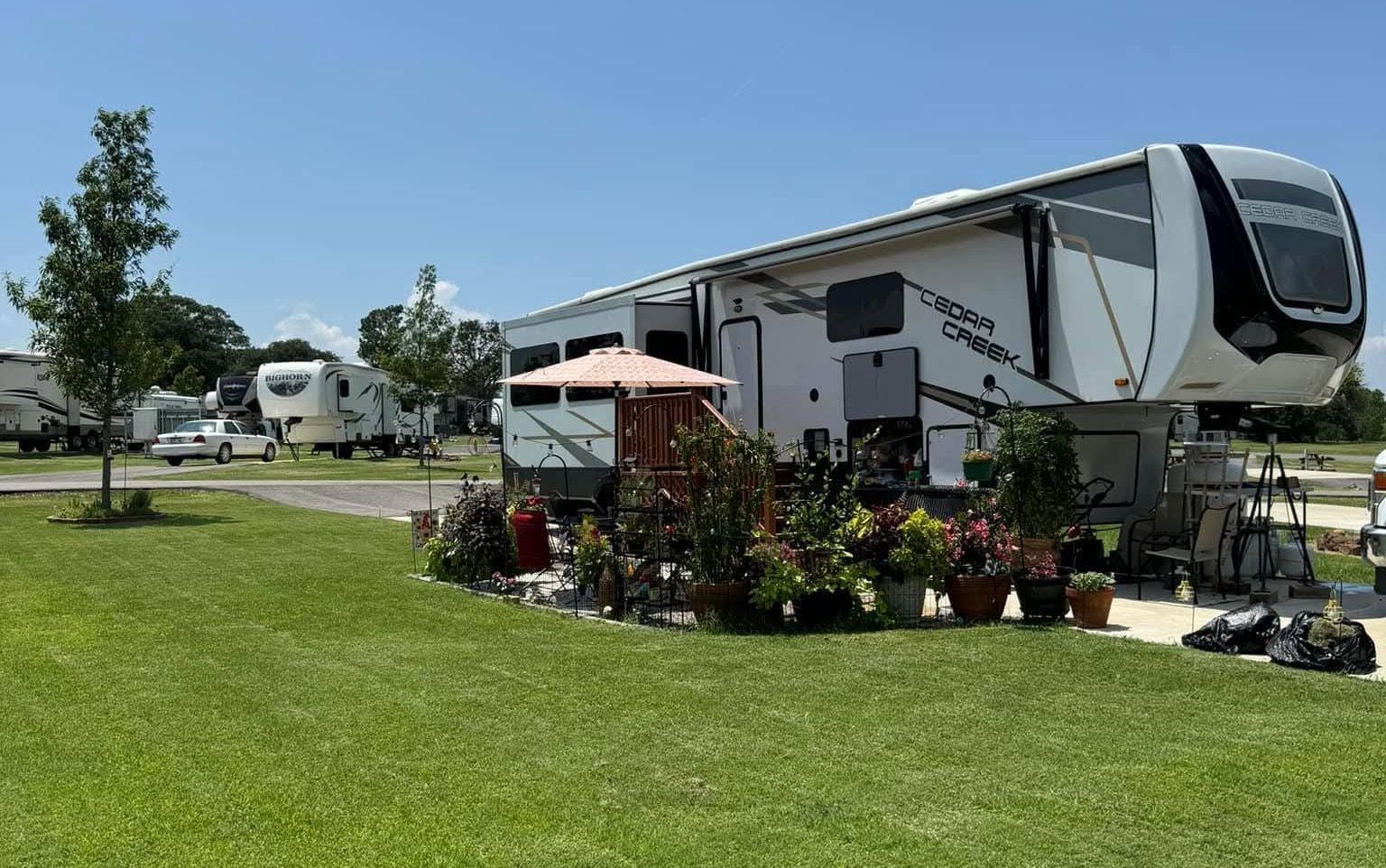 Large white RV at a sunny campground with green lawn, potted plants, and patio furniture.