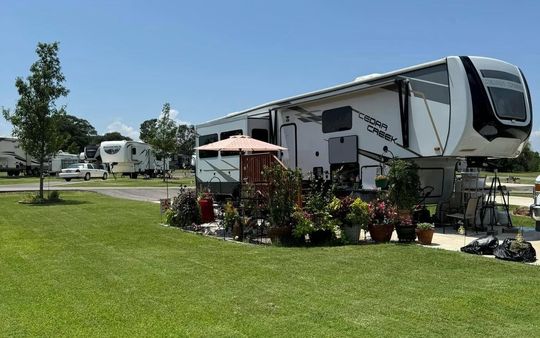 Large white RV at a sunny campground with green lawn, potted plants, and patio furniture.