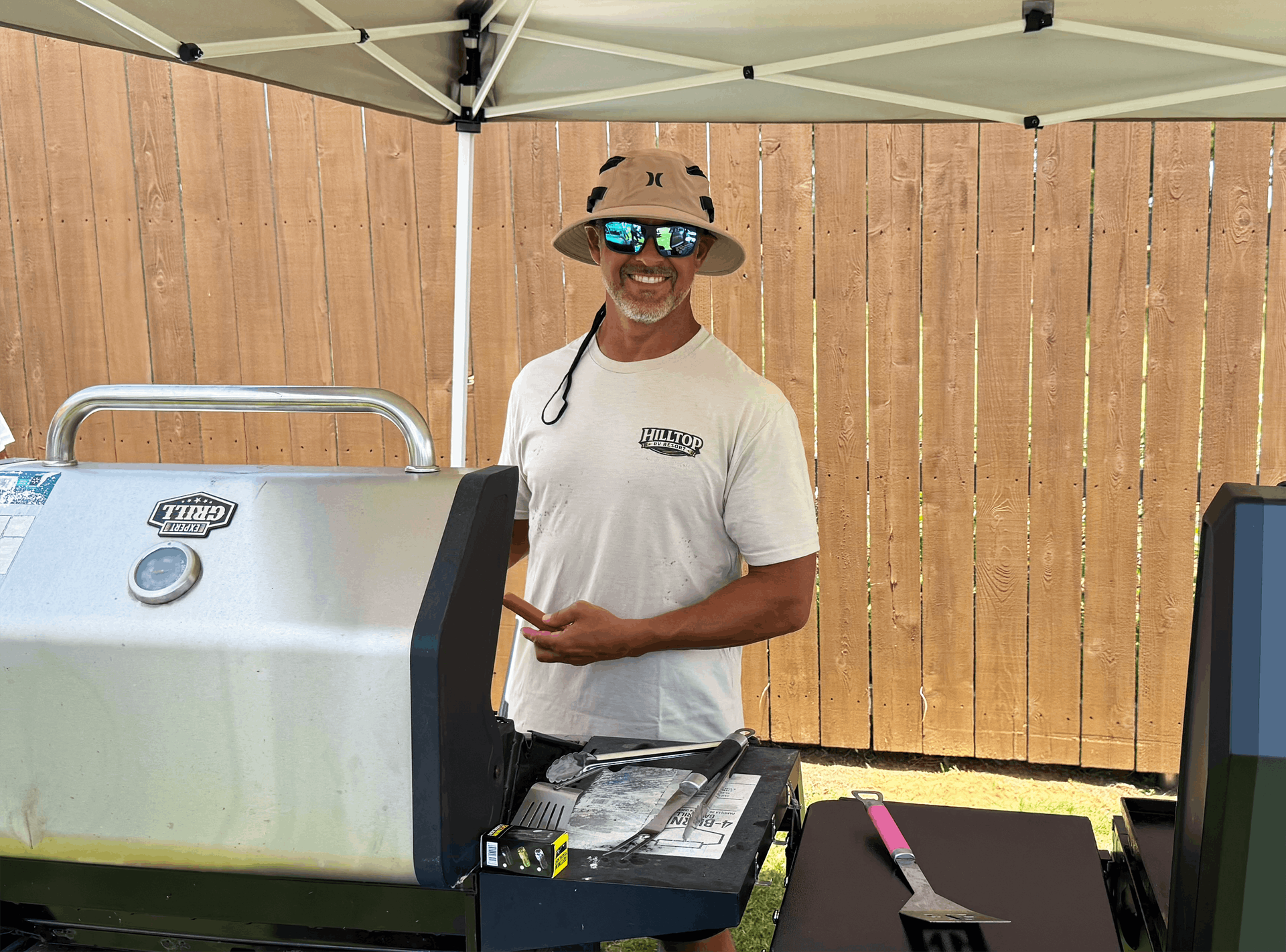 Man grilling at an outdoor barbecue under a canopy beside a wooden fence, wearing a hat and sunglasses.