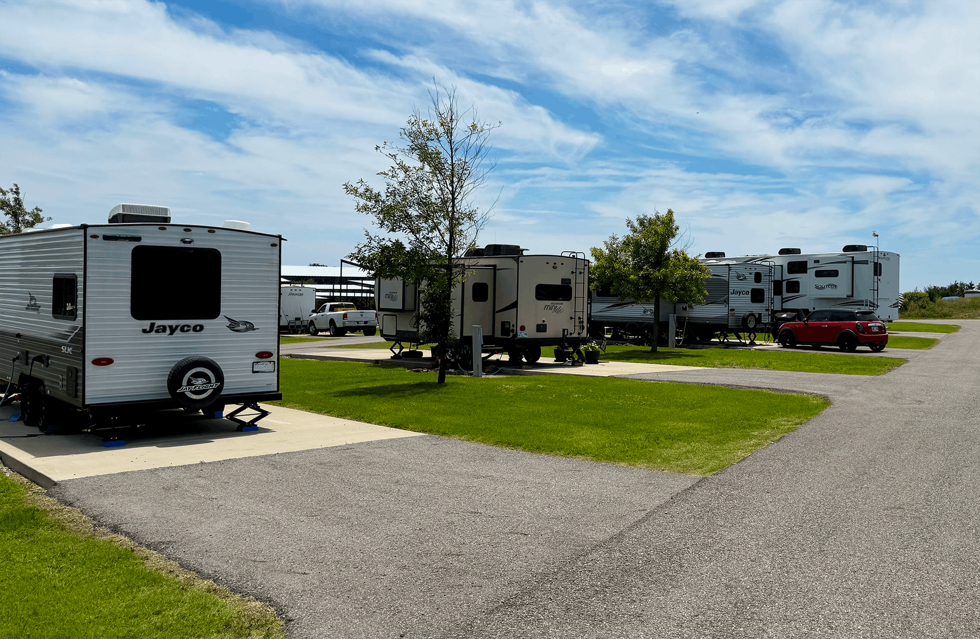 RVs parked at a campground with green grass and a paved road under a blue sky