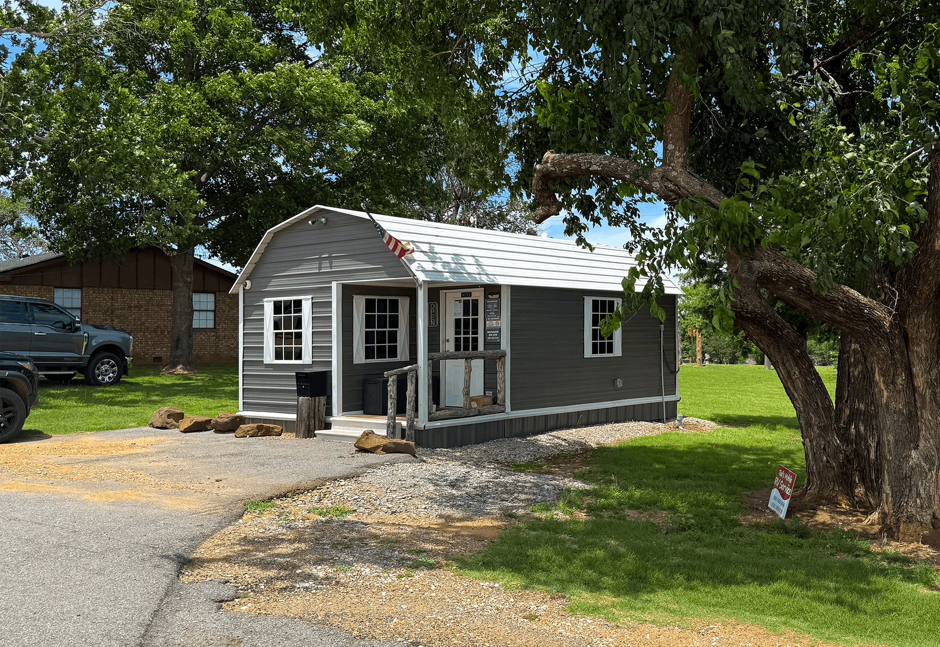 Small gray house with white trim beside a gravel driveway, shaded by large trees.