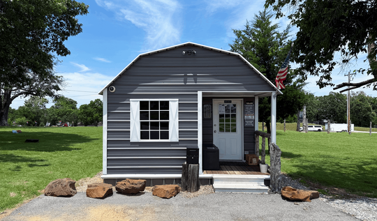 Small gray house with white trim, front porch, and rocks in a sunny grassy yard