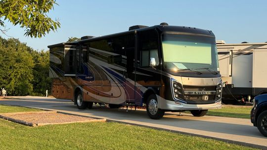 Large black-and-beige motorhome parked on a grassy roadside under trees in bright daylight