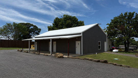 Gray barn-style building with white roof on gravel lot under blue sky, bordered by trees and grass