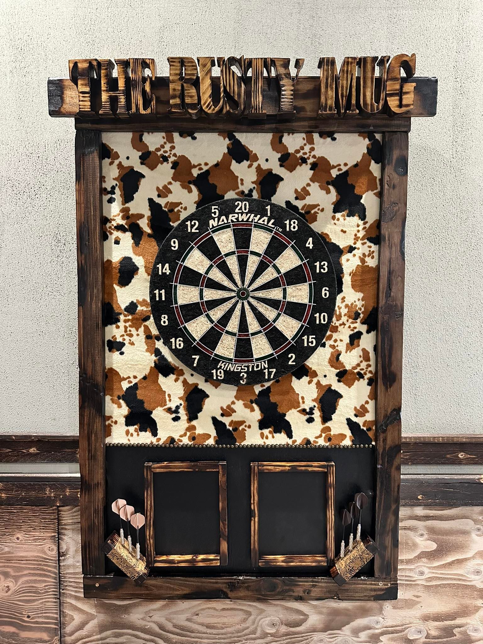 Wooden dartboard cabinet with cow print background, dartboard, and scoreboards.