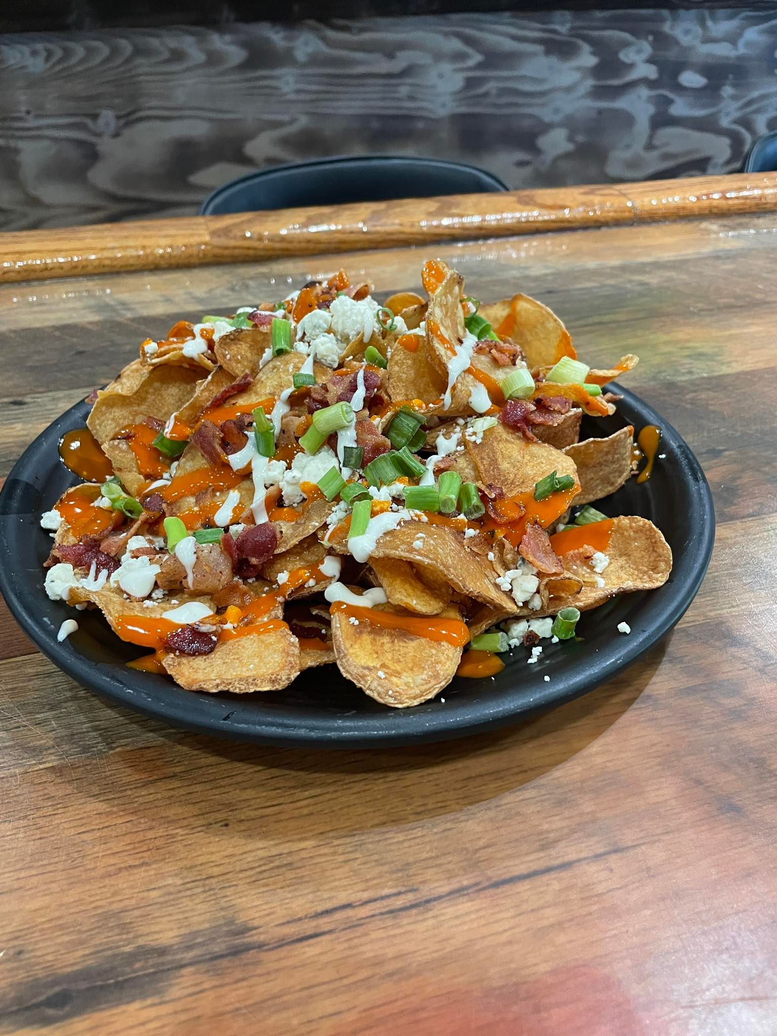 Loaded potato chips in a black bowl, topped with bacon, cheese, green onions, and sauce on a wooden table.