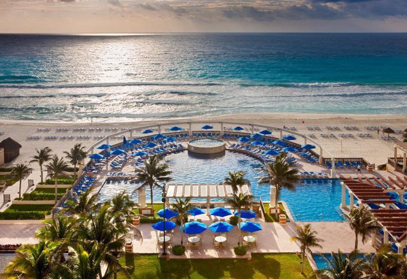 Beachfront resort pool with blue umbrellas, palm trees, and ocean view at sunset