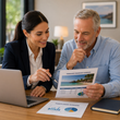 Two coworkers review charts and a beach photo at a laptop in an office meeting.