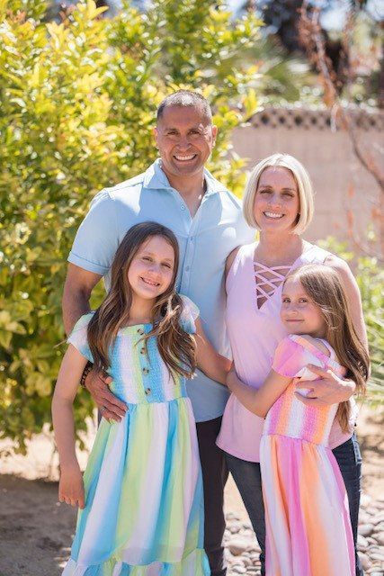 Family of four standing outdoors, smiling in bright sunlight with greenery in the background.