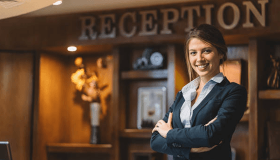 Smiling hotel receptionist in a dark blazer stands at the front desk under a “RECEPTION” sign.