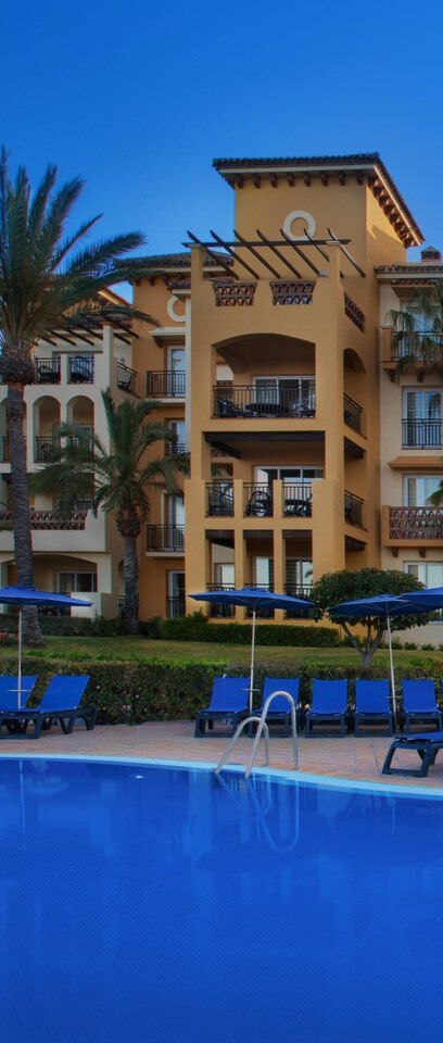 Blue hotel pool in front of a tan resort building with palm trees and lounge chairs