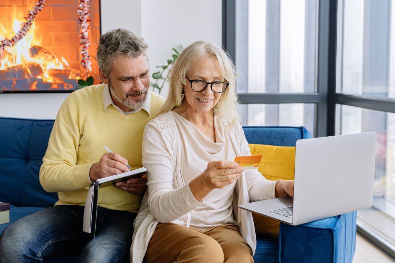 Two people on a blue couch using a laptop and tablet, holding a credit card in a bright living room.