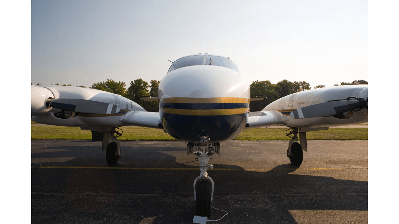 Twin-engine airplane on tarmac, front view. White, blue, and gold fuselage. Wheels down, sunny sky.