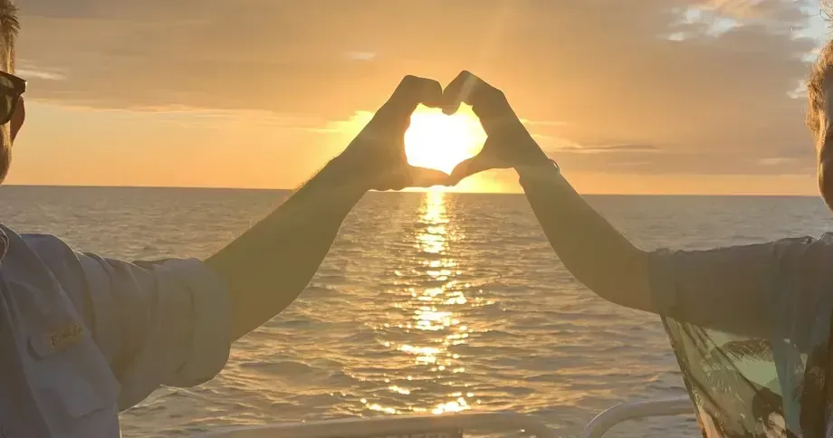 Two people form a heart with their hands at sunset over the ocean