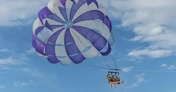 Purple-and-white parasail carrying two people against a blue sky with clouds