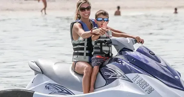 Woman and child riding a white-and-purple jet ski on calm water, both wearing life vests and sunglasses