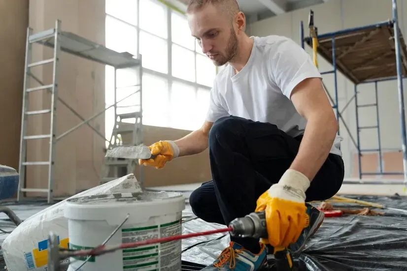 A man is kneeling down with a drill and a bucket of paint.