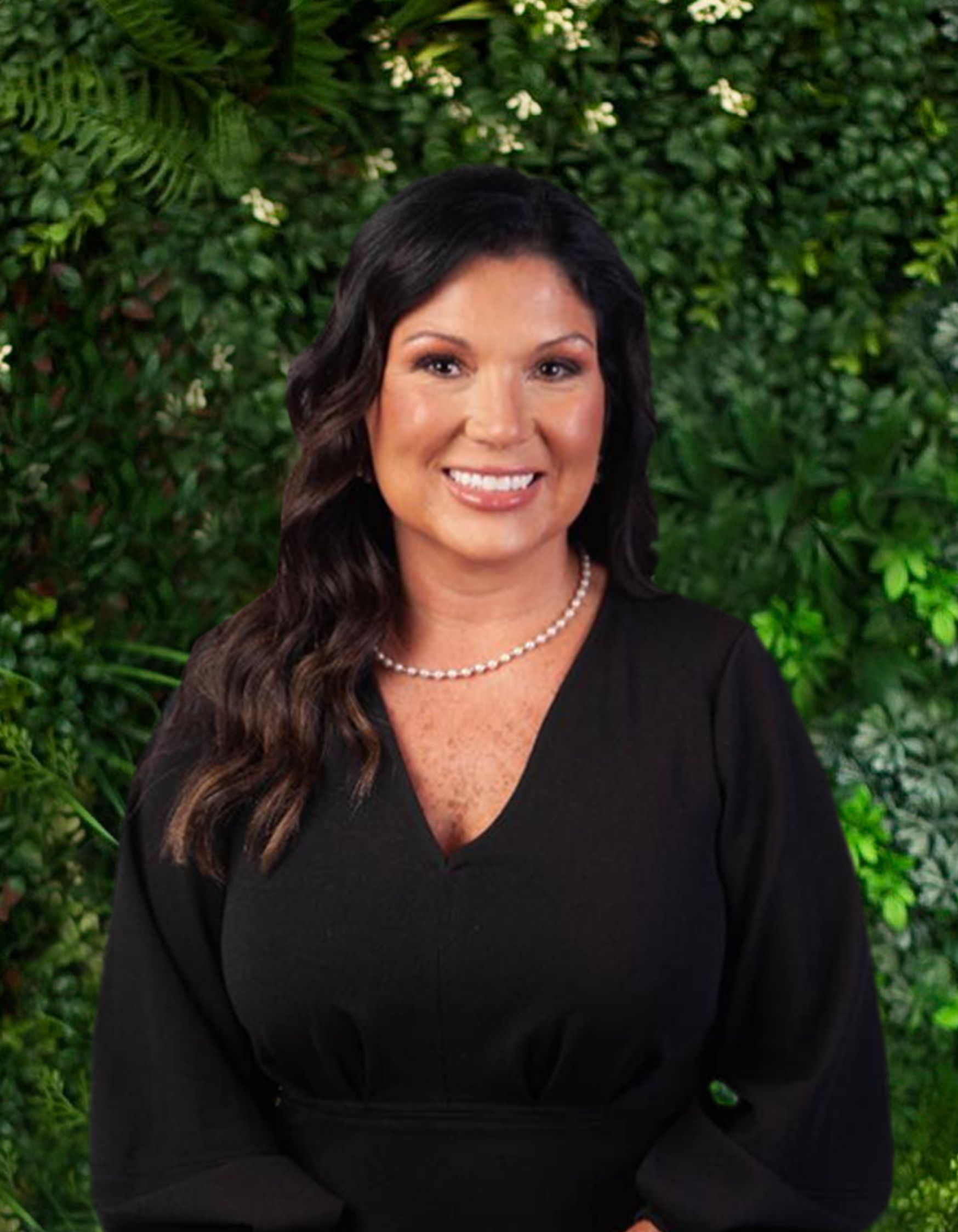 A woman in a black dress and pearl necklace is smiling in front of a green wall.
