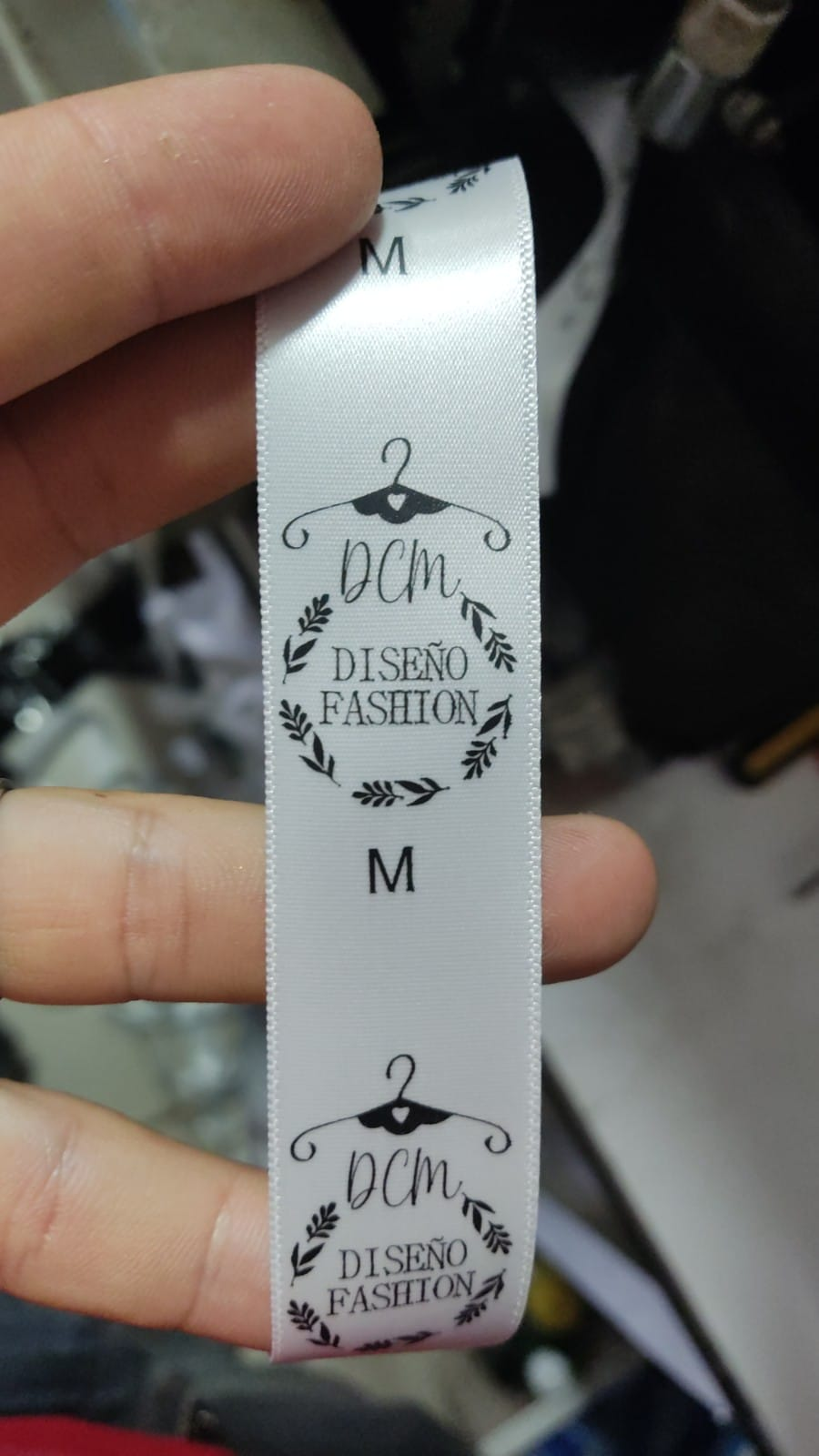 Etiqueta blanca de ropa con la talla “M” en negro y el texto “Ocm Diseno de Fashion”, sostenida entre los dedos.