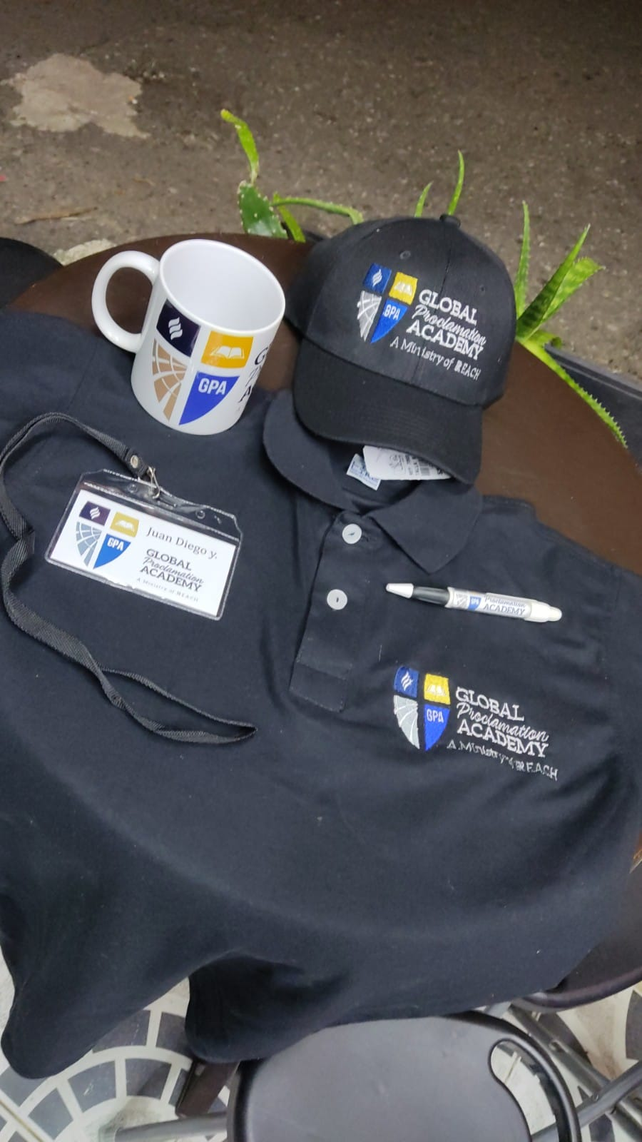 Polo negro, gorra, taza y cordón con el logo de la escuela, dispuestos sobre una mesa con una pequeña planta cerca.