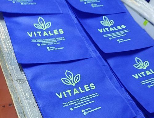 Bolsas reutilizables azules estampadas con el logotipo de VITALES dispuestas sobre una mesa.