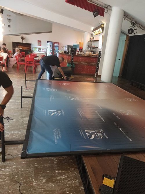 Mesa de ping-pong azul en una cafetería informal, con gente cerca y sillas rojas al fondo.