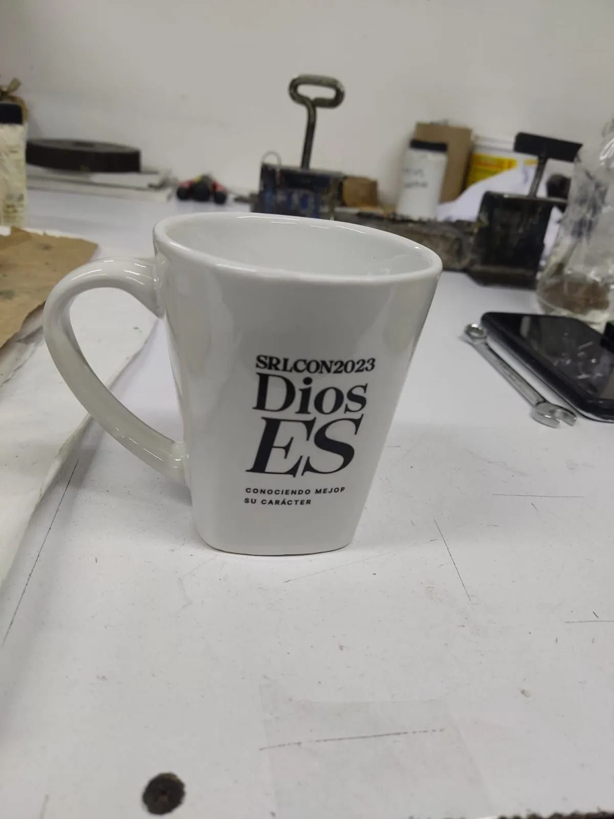 Taza blanca sobre un escritorio desordenado con texto negro en negrita que dice “Dios es”.
