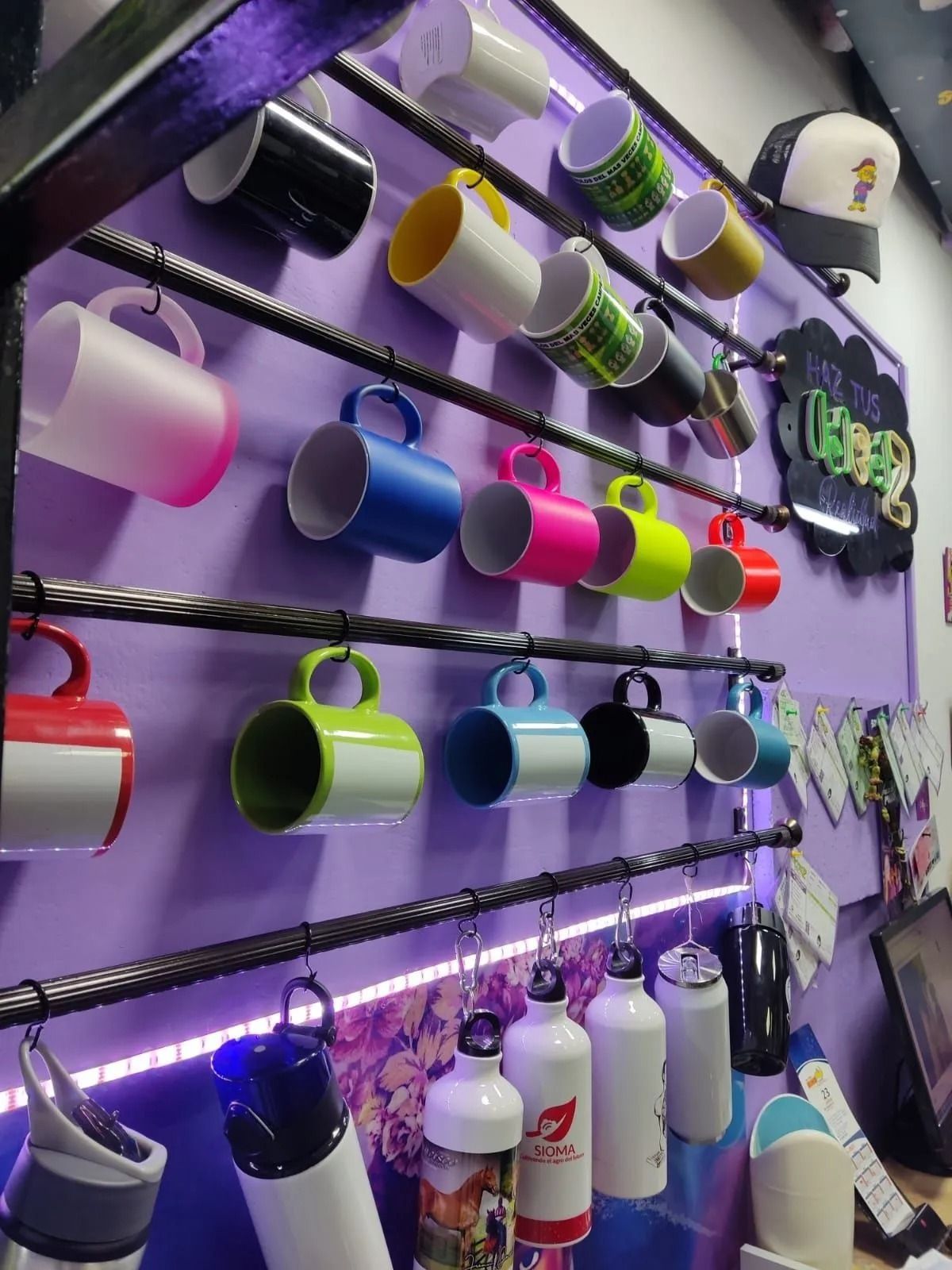 Tazas coloridas colgadas de rieles de metal negro sobre una pared morada, con botellas y accesorios debajo.