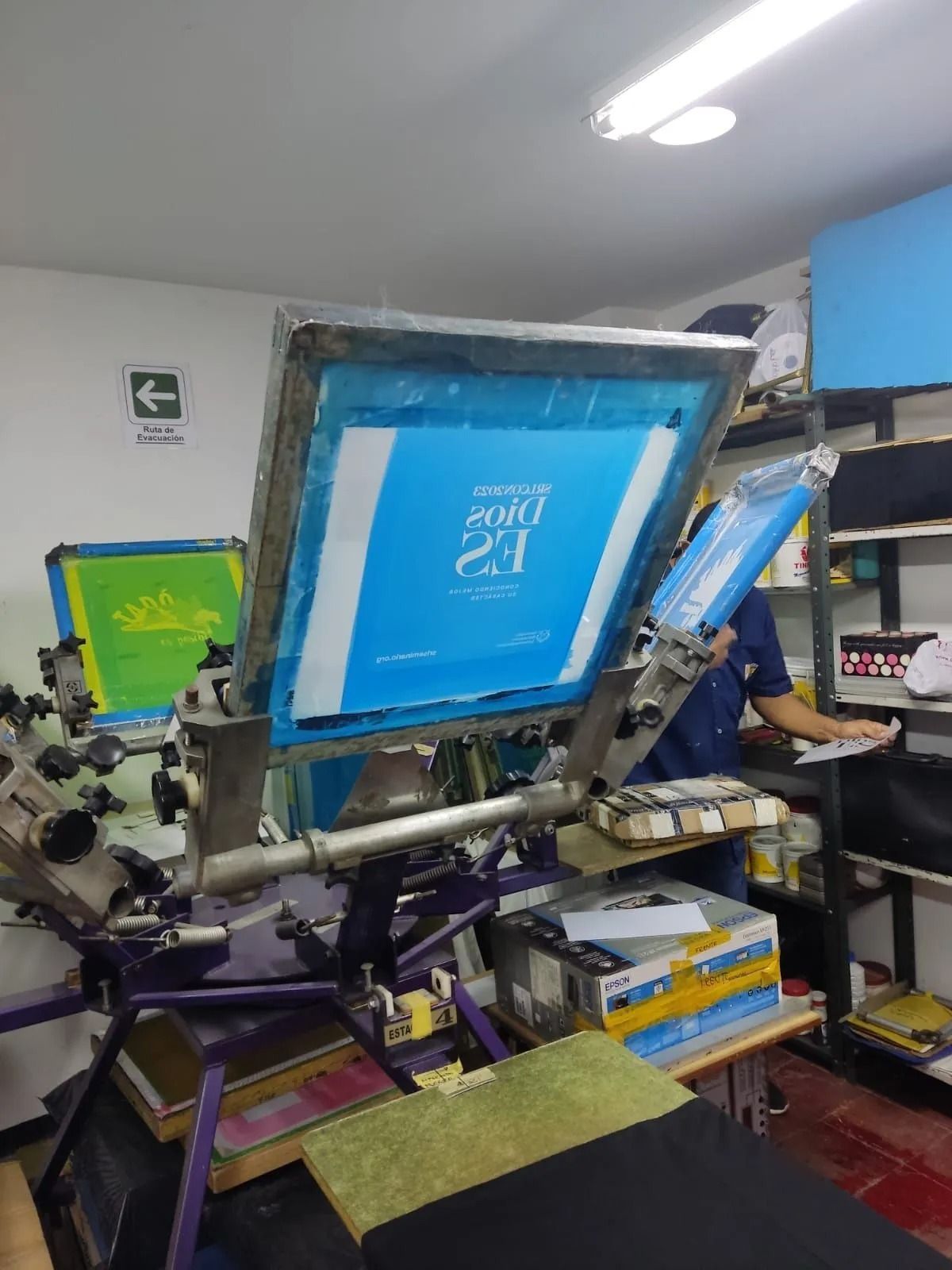 Máquina de serigrafía con pantalla azul en un taller, rodeada de herramientas y materiales.