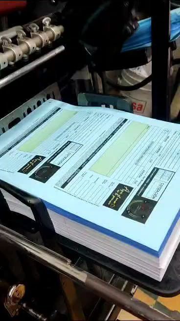 Pila de formularios impresos en la bandeja de una máquina en un entorno industrial, con los bordes del papel azul visibles.