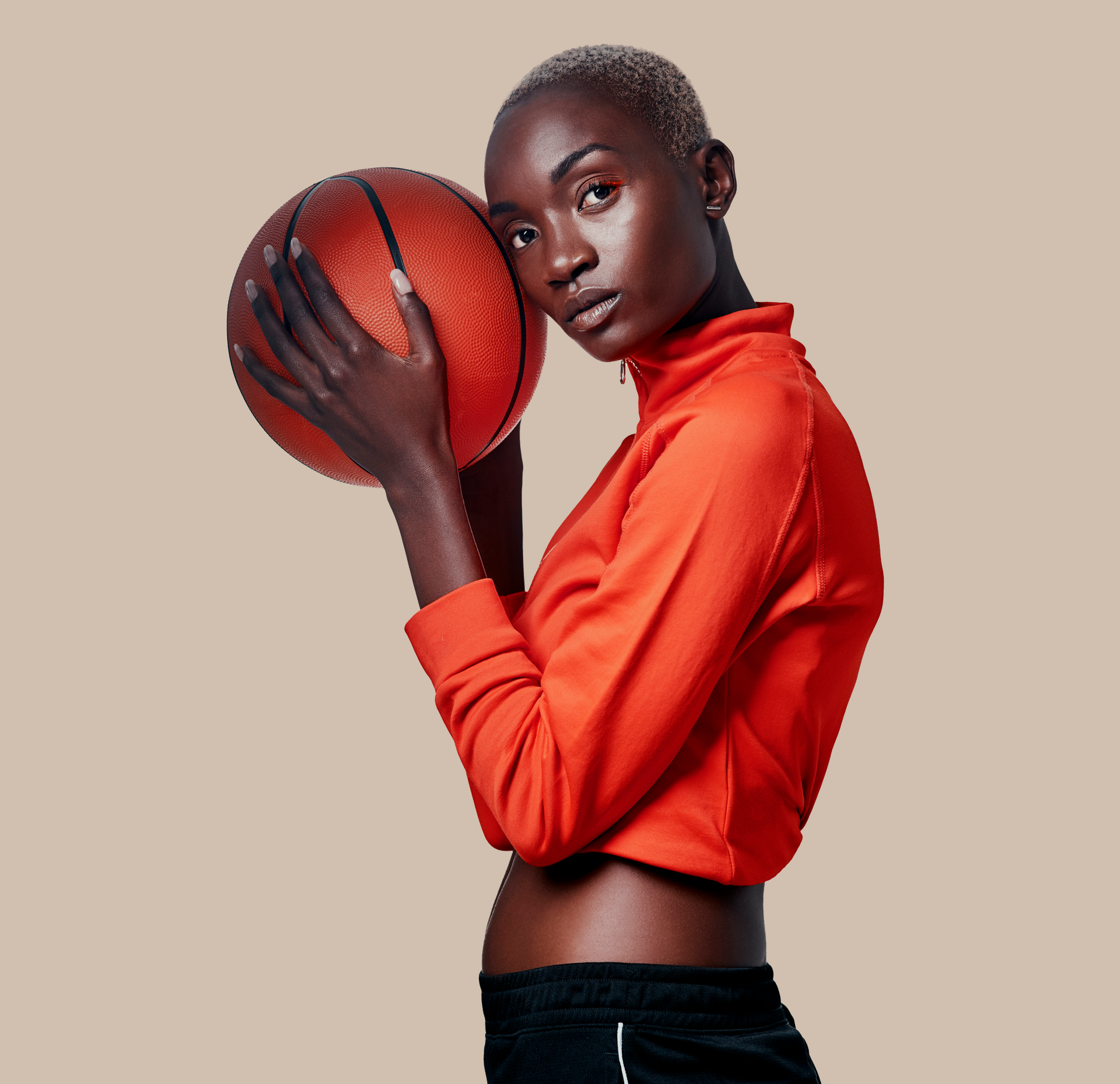 Woman with shaved head, wearing red top and black shorts, holds basketball, neutral background.