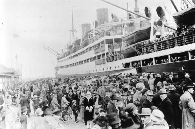 Multitud embarcando en un gran transatlántico en un muelle concurrido, con chimeneas humeantes y cubiertas abarrotadas a la vista.