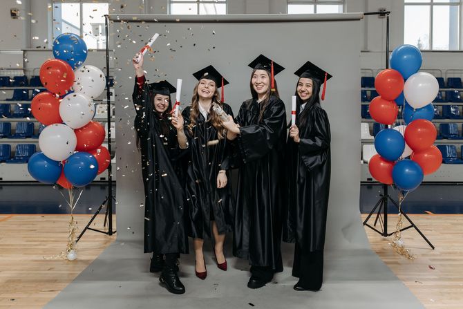 Seven people posing at a graduation celebration with balloons and a red curtain backdrop.