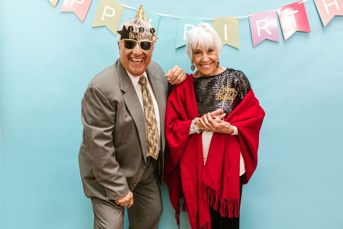 Two people dancing against a white wall, one holding a camera-like object, the other with arms raised.