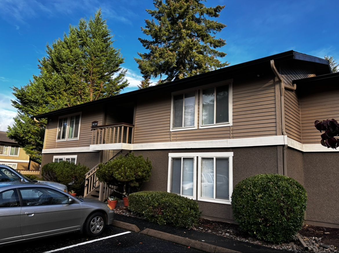Two-story apartment building with brown siding, white trim, parked cars, and trees under a clear blue sky