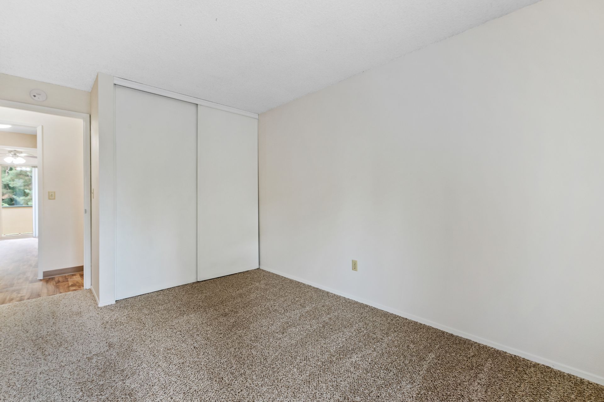 Empty beige carpeted room with white walls, closet doors, and a doorway to a hallway